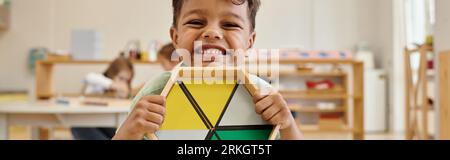 Glücklicher afroamerikanischer Junge, der Holzspiel in der verschwommenen montessori-Schule hält, Banner Stockfoto