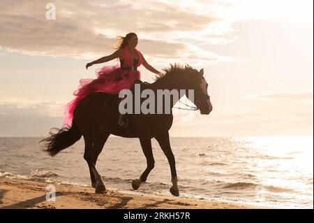 A female figure atop a majestic white horse trotting along the shoreline of a picturesque beach Stock Photo