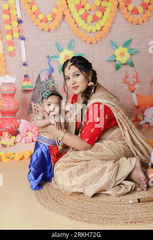 Janmashtami-Konzept. Mutter sitzt mit ihrem süßen Jungen, der als kleine krishna verkleidet ist. Stockfoto