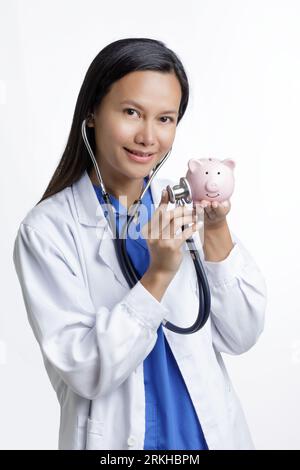 Asiatische amerikanische Ärztin untersucht eine Schweinebank, Konzept zeigen die hohen Kosten der Gesundheitsversorgung isoliert auf einem weißen Hintergrund mit Kopierraum Stockfoto