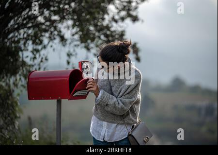 A young woman in a casual outfit stands in front of a bright red mailbox, looking inside Stockfoto