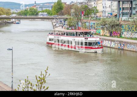 A riverboat is seen sailing down a body of water with a graffiti-covered wall and buildings along both sides of the shore in Vienna, Austria Stock Photo