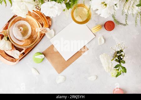 Beige crafting paper envelope with stationery card for greetings, invitation, text among white flowers, macaroons and tea. Biophilic interior, cozy ho Stock Photo