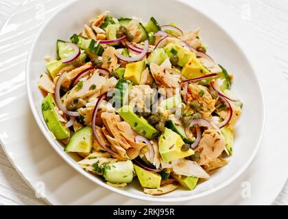 Thunfischsalat mit Avocado, roter Zwiebel, Gurke und Kapern mit Olivenöl-Limetten-Petersilie-Dressing in weißer Schüssel auf weißem Holztisch Stockfoto