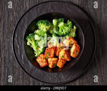baked salmon bites with steamed broccoli florets in black bowl on dark wooden table with sriracha mayo sauce, horizontal view from above, close-up Stockfoto