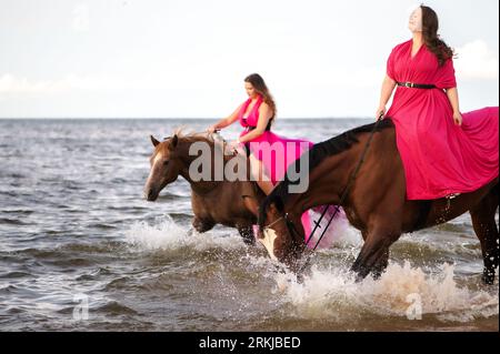 Die beiden Reiterinnen in leuchtend rosa Kleidern führen zwei majestätische Pferde entlang einer Strandküste. Stockfoto