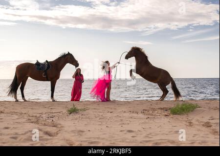 Die beiden Reiterinnen in leuchtend rosa Kleidern führen zwei majestätische Pferde entlang einer Strandküste. Stockfoto