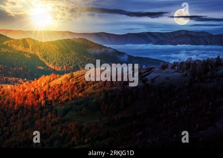 Kalter Nebel im ländlichen Tal der Karpaten mit Sonne und Mond in der Dämmerung. Bäume mit buntem Laub auf der Hangwiese am Morgen Stockfoto