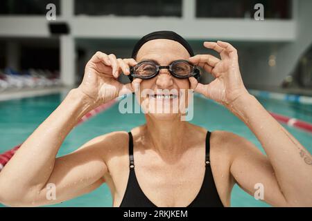 happy and tattooed woman in swim cap wearing goggles, positive, swimming pool, recreation center Stockfoto