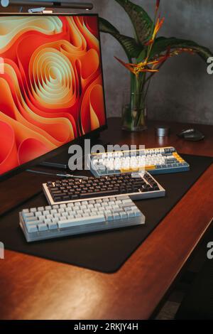 A computer with three different mechanical keyboards and a mouse lying on the desk next to the monitor Stock Photo