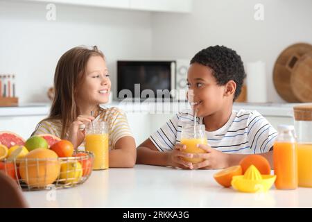 Little children with glasses of fresh citrus juice sitting at table in kitchen Stock Photo