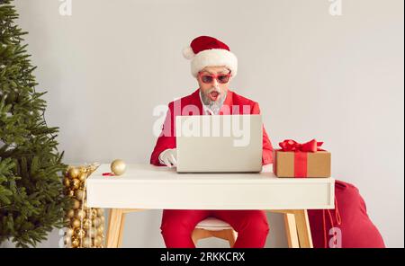 Der alte, verblüffte Mann in rotem Anzug, Weihnachtsmannmütze, Sonnenbrille, die am Schreibtisch sitzt und am Laptop in der Nähe des Weihnachtsbaums arbeitet. Stockfoto