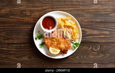 Hausgemachtes paniertes wiener Schnitzel mit Kartoffelfrites und Sauce auf weißem Teller auf hölzernem Hintergrund mit freiem Platz. Draufsicht, flach Stockfoto