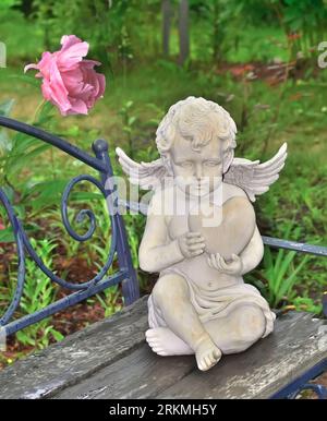 Gemütliche, romantische Gartenecke mit Engelskuppel auf Holzbank mit Herz in den Händen. Schöne rosa Pfingstrosenblüte, die über seinem Kopf blüht. Summ Stockfoto