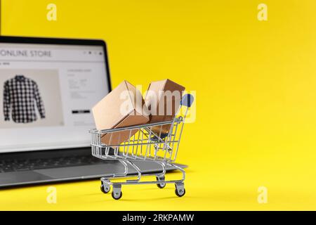 Online-Shop. Mini-Warenkorb, Pakete und Laptop auf gelbem Hintergrund, selektiver Fokus. Leerzeichen für Text Stockfoto