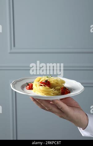 Kellner hält Teller mit leckeren Capellini mit Tomaten und Käse nahe der grauen Wand, Nahaufnahme und Platz für Text. Exquisite Präsentation von Nudelgerichten Stockfoto