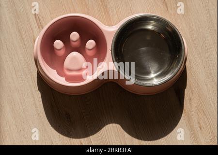 Eine Doppelschüssel für langsames Füttern und eine Schüssel Wasser für den Hund. Draufsicht auf einen rosafarbenen Teller mit trockenen Speisen auf einem Holzboden. Stockfoto