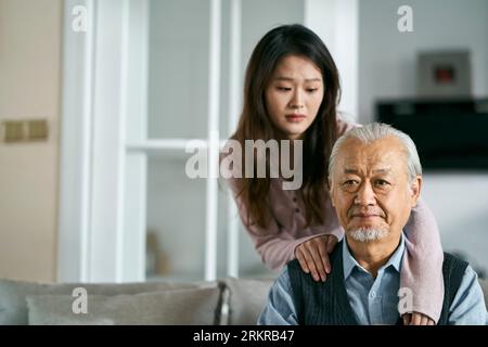 Trauriger älterer asiatischer Vater, der auf der Couch im Wohnzimmer zu Hause saß, getröstet von einer erwachsenen Tochter Stockfoto