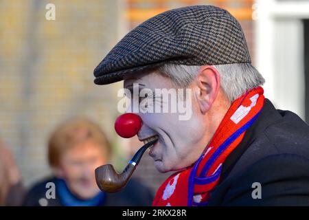 Porträt eines Mannes mit einem blassweißen Gesicht, einer Clownsnase, einer Pfeife im Mund mit einer Kappe und einem rot-blauen Schal um den Hals: Karneval Stockfoto