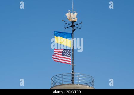 Die Flaggen der Ukraine und der USA flattern auf einem Fahnenmast vor einem blauen Himmel. Statussymbol. Ukrainische blaue und gelbe Flagge. Windtag im Freien. Stockfoto