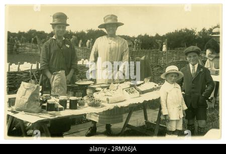 Ursprüngliche Postkarte aus der frühen 1900er-Jahre mit einem Bring-and-Buy-Stand. Neben ihrem Stall steht eine Dame mit einem Standhalter, der Stall zeigt Marmeladen, Fruchtnougats, Dämonen am Pfund - Waagen im Hintergrund. Zwei junge Kunden stehen neben dem mit Waren zum Verkauf ausgelegten Stehtisch. Der Junge trägt einen HMS Defender Matrosenhut und das andere Kind eine flache Kappe. CA. 1919 /1920, Großbritannien Stockfoto