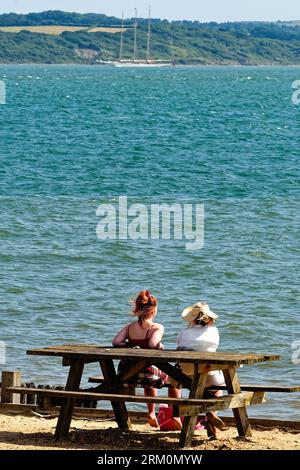 Zwei junge Frauen sitzen auf einer Tischbank am Strand von Lepe und blicken an einem heißen Sommertag über den Solent zur Isle of Wight in Hampshire England Stockfoto