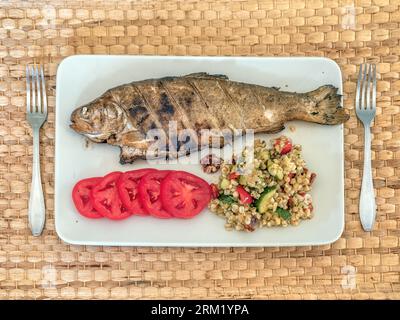 Gegrillte ganze Forelle serviert auf einem Teller mit Tomatenscheiben und Grobgrieß Stockfoto