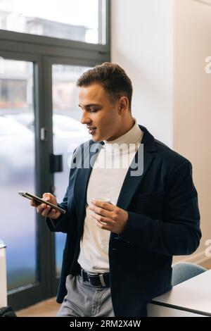 Ein junger, stilvoller Geschäftsmann schaut sich sein Telefon an, während er den ersten Kaffee für den Tag, früh am Morgen, trinkt Stockfoto