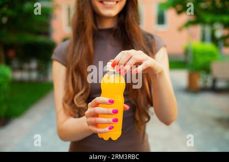 Eine nicht erkennbare junge Frau öffnet an einem Sommertag draußen eine orange Limonade-Plastikflasche, die in einer Stadtstraße in bunten Gebäuden steht Stockfoto