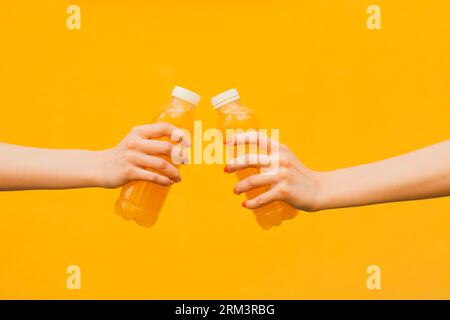 Zwei nicht erkennbare junge Frauen halten in seiner Hand die orange Limonade Plastikflasche auf gelbem Straßenmauerhintergrund, Nahaufnahme Seitenansicht Stockfoto