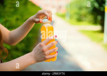Eine nicht erkennbare junge Frau öffnet an einem Sommertag draußen eine orange Limonade-Plastikflasche, die in einer Stadtstraße in bunten Gebäuden steht Stockfoto