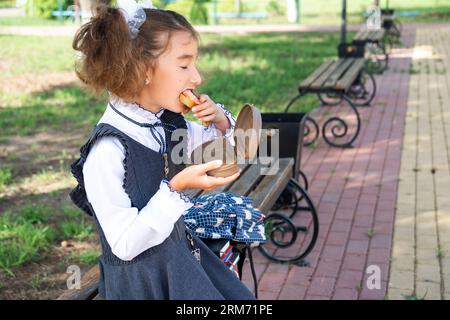 Mädchen mit Rucksack, das Sandwich in einer Sandwichbox in der Nähe der Schule isst. Ein schneller Snack mit einem Brötchen, ungesundes Essen, Mittagessen aus der Schule. Zurück zur Schule. Stockfoto