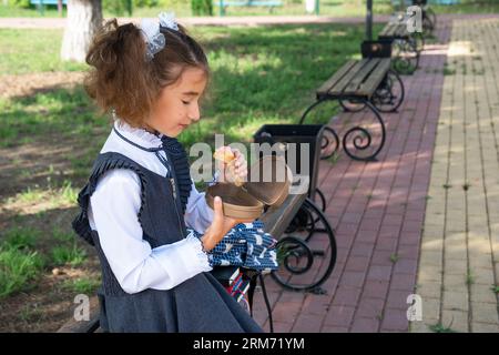 Mädchen mit Rucksack, das Sandwich in einer Sandwichbox in der Nähe der Schule isst. Ein schneller Snack mit einem Brötchen, ungesundes Essen, Mittagessen aus der Schule. Zurück zur Schule. Stockfoto