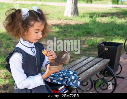 Mädchen mit Rucksack, das Sandwich in einer Sandwichbox in der Nähe der Schule isst. Ein schneller Snack mit einem Brötchen, ungesundes Essen, Mittagessen aus der Schule. Zurück zur Schule. Stockfoto