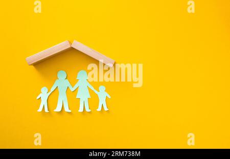 Papierausschnitte einer Familie, die unter einem Holzdach auf einem gelben Hintergrund stehen und Heimschutz symbolisieren Stockfoto