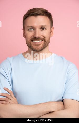 Glücklicher Mann weiße Zähne, die mit gekreuzten Armen lächeln, schauen auf die Kamera, die isoliert auf der rosa Studiowand steht Stockfoto