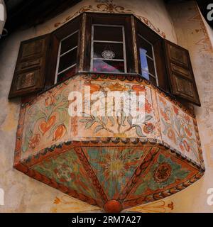 Traditionelles Balcun Tort- oder Erkerfenster mit verzierten Holzläden und lebendiger Blumen- und Vogeldekoration. Auf einem traditionellen Haus in Guarda, einem Dorf im unteren Engadintal in Graubünden oder Bündner Kanton. Balcun Tort ist ein romanischer Begriff und diese Fenster sind typisch für die romanisch sprechende Schweiz. Zwillings- und Dreieckserker sind bei älteren Häusern im Kanton Graubünden üblich. Stockfoto