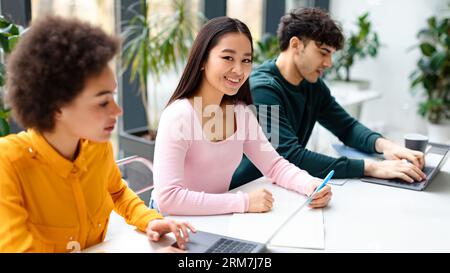 Studienprozess. Verschiedene Klassenkameraden sitzen am Schreibtisch, asiatische Dame lächelt vor der Kamera, während sie sich auf die Hochschulprüfung vorbereitet Stockfoto