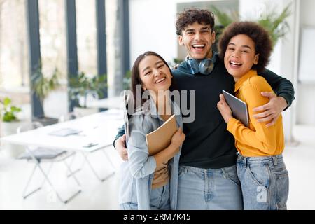 Drei begeisterte Freunde von internationalen Studenten, die sich in die Kamera umarmen und lächeln, sich im Klassenzimmer posieren, im Kopierraum Stockfoto