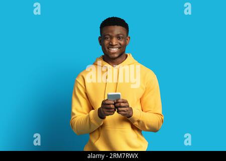Glücklicher junger schwarzer Mann in gelbem Hoodie mit Smartphone Stockfoto