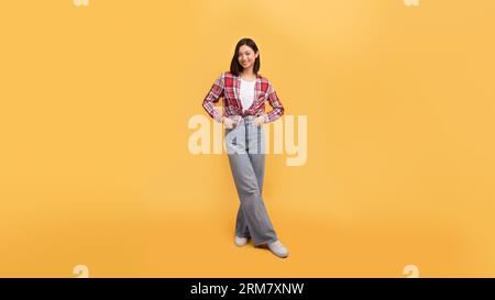 Aufnahme einer glücklichen Dame in lässigem Outfit in voller Länge, die Hände in den Taschen hält und vor der Kamera lächelt, gelber Hintergrund Stockfoto