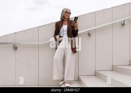 Glückliche reife alte Frau mit grauen Haaren und Brille trägt eine stilvolle Jacke und geht zu einem Geschäftstreffen Stockfoto