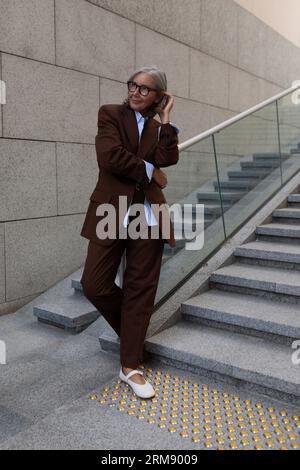 Die charmante, erfolgreiche Seniorin mit grauen Haaren und Brille trägt einen braunen Designer-Anzug Stockfoto