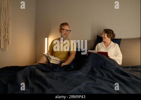 Happy laughing senior couple lying in bed with book and talking Stockfoto