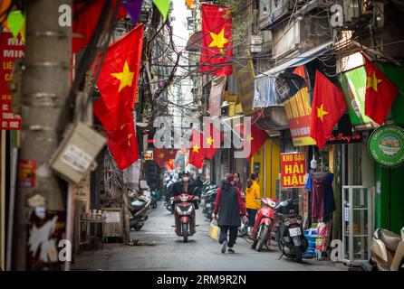 Die Menschen gehen unter vietnamesischen Flaggen in einer engen Gasse namens Kham Thien Market in Hanoi, Vietnam, über das tägliche Leben. Stockfoto