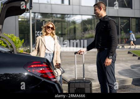 Businesswoman waits while chauffeur packs a suitcase in car trunk Stockfoto