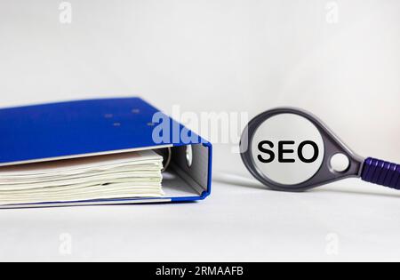 Das Wort SEO steht auf einem Lupenglas und einem weißen Hintergrund mit einem blauen Dokumentenordner Stockfoto