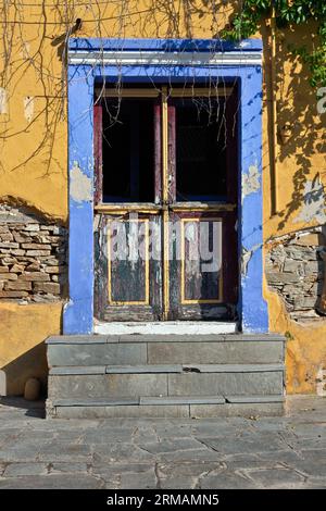 Wunderschöner Eingang mit alter Holztür, blauem bis violettem Sturz, gelber Wand und Zementtreppe bei warmem Nachmittagslicht, in der Stadt Eleftheroupoli Stockfoto