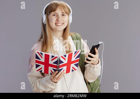 Lächelndes, trendiges Schulmädchen im beigefarbenen Trainingsanzug mit Rucksack, Arbeitsbuch mit englischer Flagge, Kopfhörer und Smartphone isoliert auf grauem Hintergrund. Stockfoto