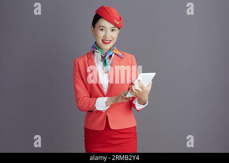 Lächelnde, stilvolle, asiatische Gastgeberin in rotem Rock, Jacke und Hut Uniform mit Tablet-PC vor grauem Hintergrund. Stockfoto